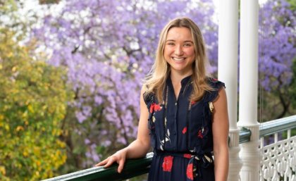 A young woman wearing a navy floral dress stands holding a railing with a flowering jacaranda tree in the background. She has blonde hair and is smiling.